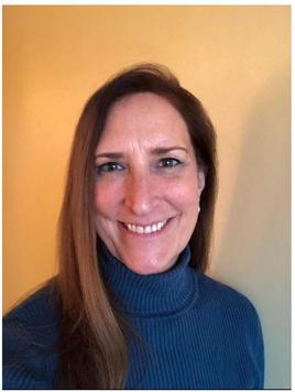 Headshot of Denver manual therapist Gigi Degerlund Willett, also known as artist Gigi Degerlund, smiling, with long reddish brown hair, wearing a blue turtleneck, in front of a yellow wall